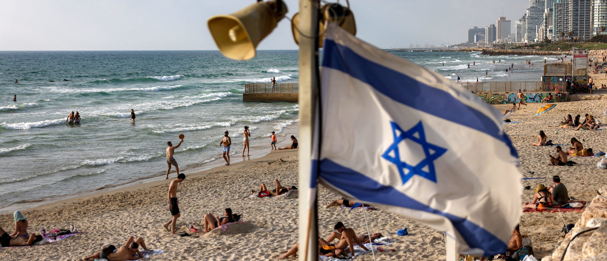 An Israeli flag flies beneath siren loudspeakers as people gather by the surf at a beach in Tel Aviv on June 21, 2025. (Photo by AHMAD GHARABLI/AFP via Getty Images)
