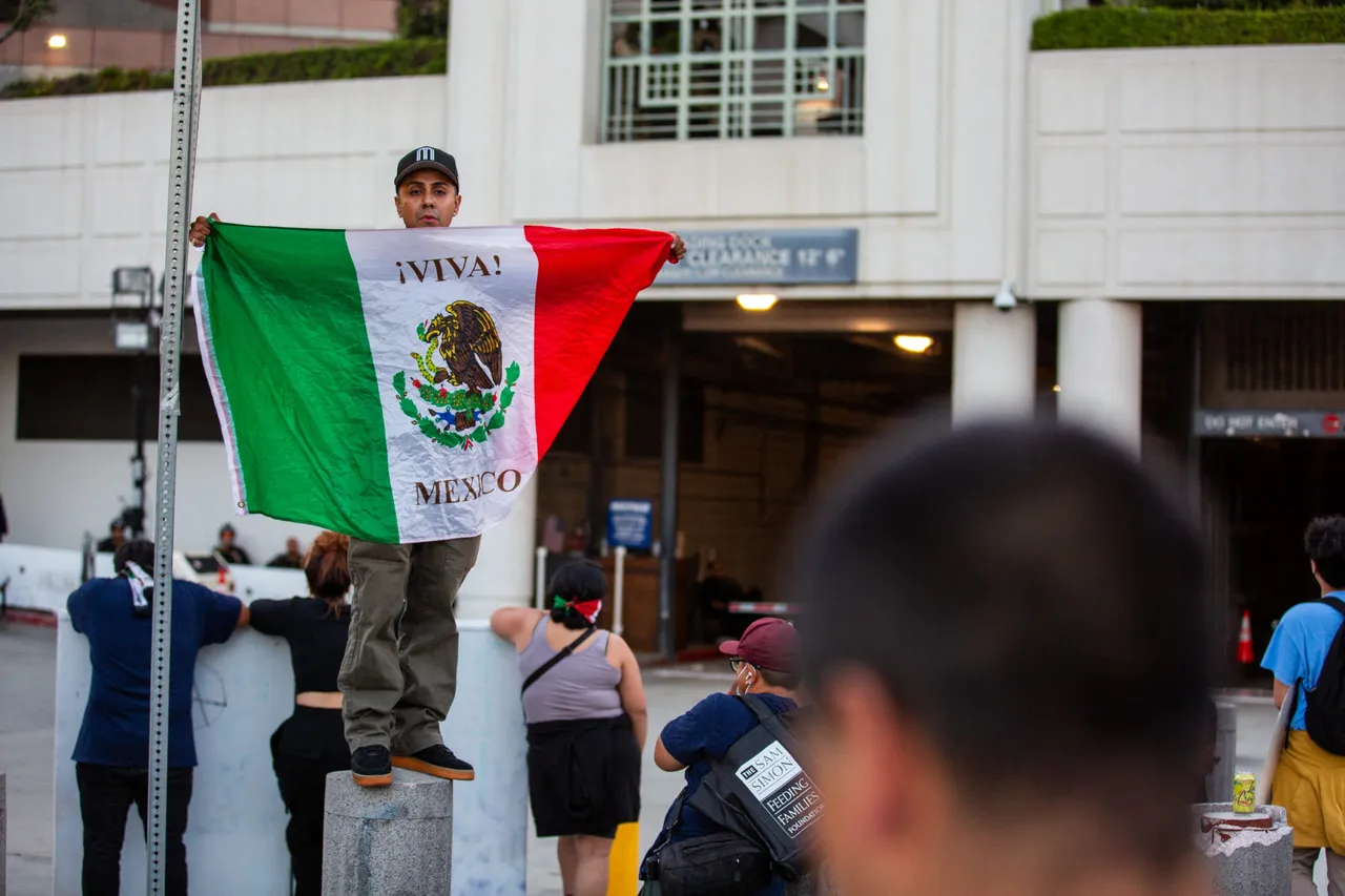 A protester waves a Mexican flag above a crowd during an Anti-ICE protest. (Photo by BENJAMIN HANSON/Middle East Images/AFP via Getty Images)